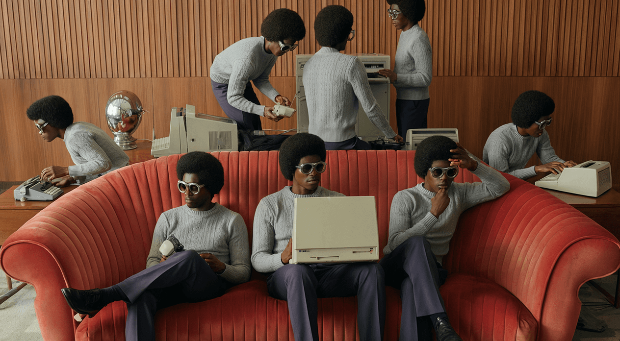 Artistic image of multiple identical men in gray sweaters, navy pants, and sunglasses working with vintage computers and typewriters around a retro red sofa.